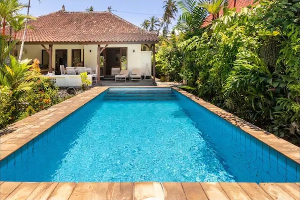 A rectangular swimming pool with a wooden deck, surrounded by lush greenery and a house with a tiled roof.