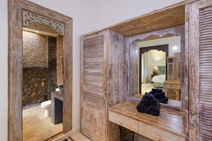A bathroom with a stone wall, wooden vanity, and a mirror reflecting a room with a bed and a lamp.