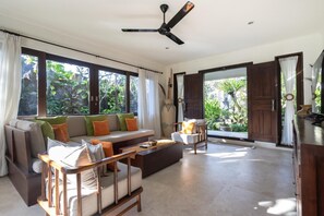 A spacious living area with a sofa, coffee table, and a chair. There is a large window with curtains, a ceiling fan, and a view of greenery outside.