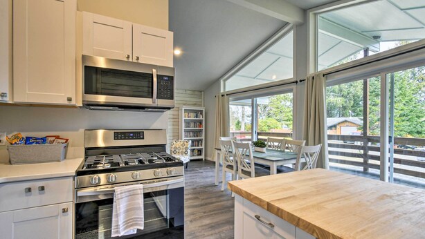 Cocina moderna con muebles blancos, horno empotrado, microondas y estufa a gas. Cuenta con un comedor con mesa y sillas, y ventanas grandes que ofrecen vistas al exterior.