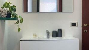 Un baño con un lavamanos blanco, un espejo encima, una planta en maceta sobre una estantería y una puerta con cerradura.