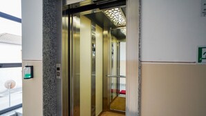 Interior de un ascensor con piso alfombrado, dispensador de gel antibacterial y puerta de vidrio.