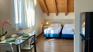 Habitación compacta con dos camas, una zona de comedor con mesa y sillas, una pequeña cocina y un techo con vigas de madera a la vista.