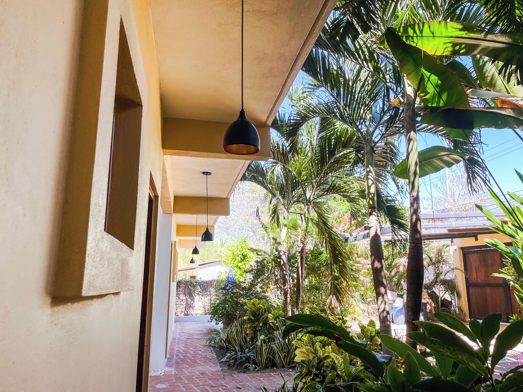 A covered outdoor area with hanging pendant lights, a brick pathway, and lush tropical plants.