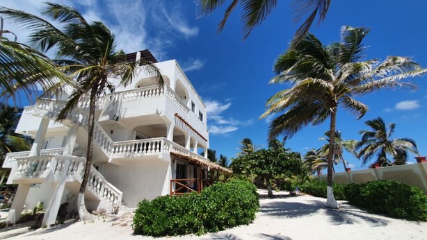 A white multi-story hotel with balconies, surrounded by palm trees and a sandy beach. - Permit- Beachfront Apartment (6 guests), Level 1 (Xcalak)