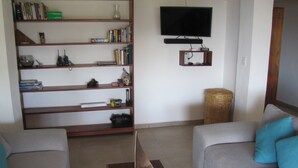 A living room with a bookshelf, a television mounted on the wall, and two grey sofas with blue cushions. - Permit- Beachfront Apartment (6 guests), Level 1 (Xcalak)