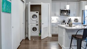 Ett modernt kök med inbyggd tvättmaskin och torktumlare, vita skåp, en mikrovågsugn och en bardisk med en diskho och barstolar.