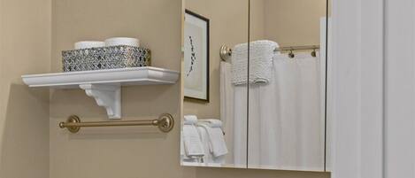 Un baño con inodoro blanco, lavamanos con encimera de mármol, tocador con armarios de madera, toallero y espejo con luces arriba.
