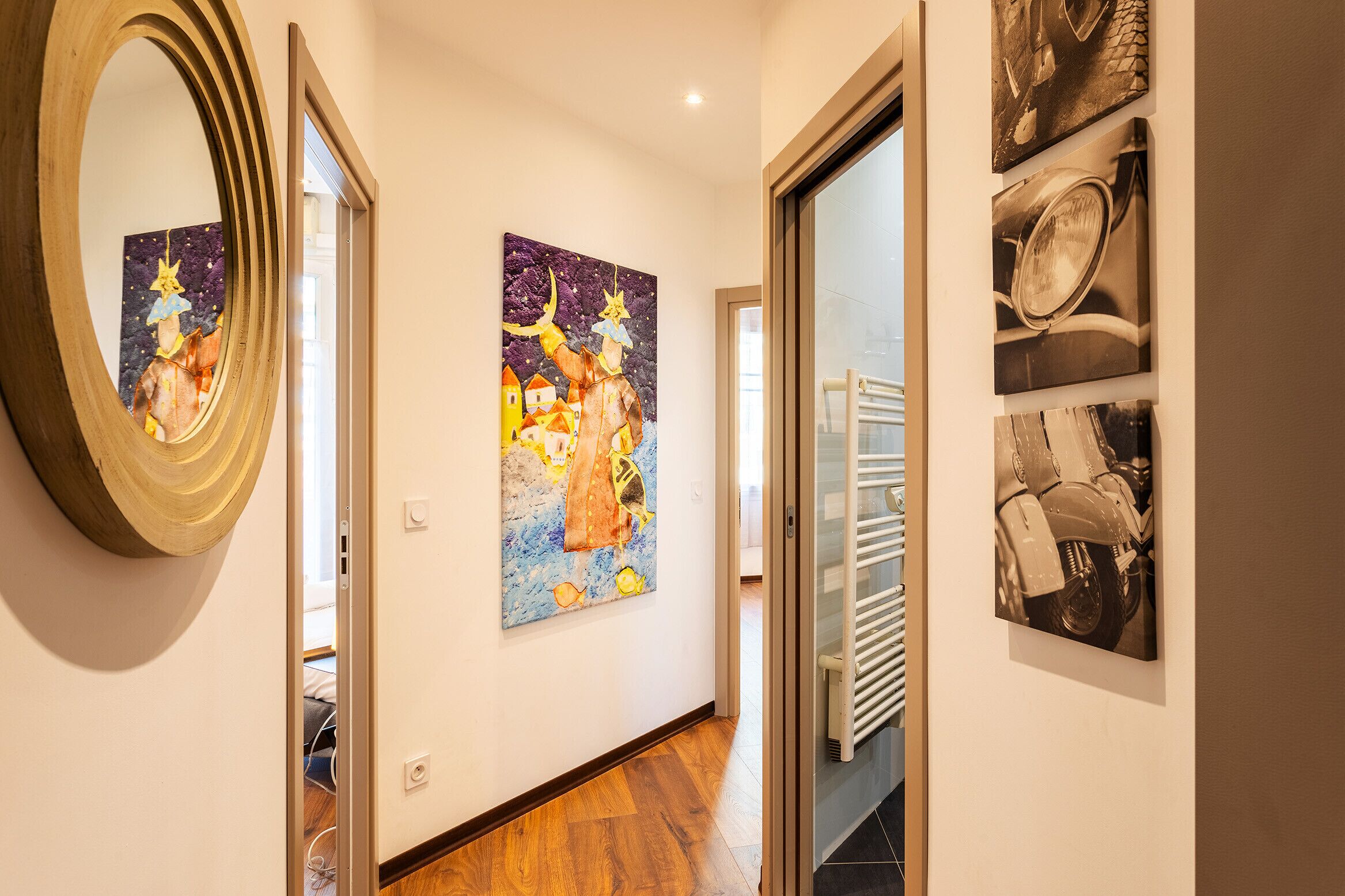 A hallway with a wooden floor, a mirror, and two paintings on the wall.