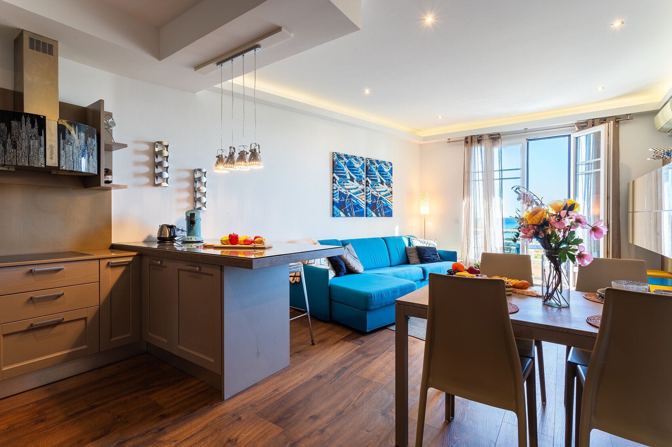 A modern kitchen with a central island, a dining area with a table and chairs, a blue sofa, and a large window with curtains.