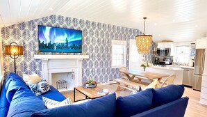 Ein modernes Wohnzimmer mit einem blauen Sofa, einem Kamin, einem Fernseher und einer Küche im Hintergrund.