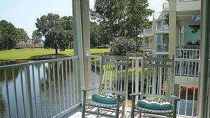 A wooden deck with two green cushioned chairs and a small table, overlooking a body of water and a well-maintained lawn with trees. - 805 2 Bedroom Villa by RedAwning (Calabash)