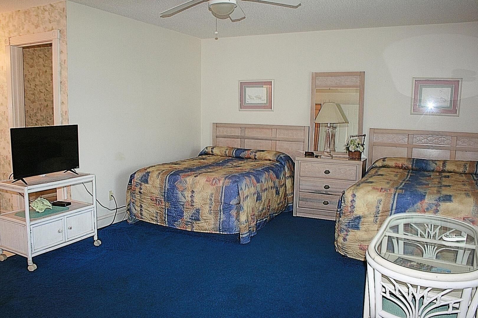 Habitación de hotel con dos camas, un televisor sobre un soporte, un aparador y un ventilador de techo.