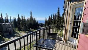 A balcony with a glass table and two wicker chairs, overlooking a forested area and a distant view. - Ski in/ski out chalet. Mountain + valley views. Close to village. (Silver Star)
