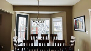 A dining room with a wooden table and chairs, a chandelier, and a view of a snowy neighborhood through large windows. - Ski in/ski out chalet. Mountain + valley views. Close to village. (Silver Star)