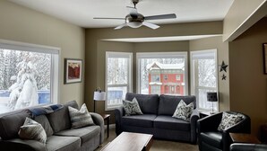 A living room with two sofas, a coffee table, and a ceiling fan. There are large windows with views of a snowy landscape and a building. - Ski in/ski out chalet. Mountain + valley views. Close to village. (Silver Star)