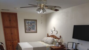 Een gemeubileerde woonkamer met een bank, een houten salontafel, een bed met hoofdbord, een bureau met stoel, een televisietoestel en een plafondventilator.