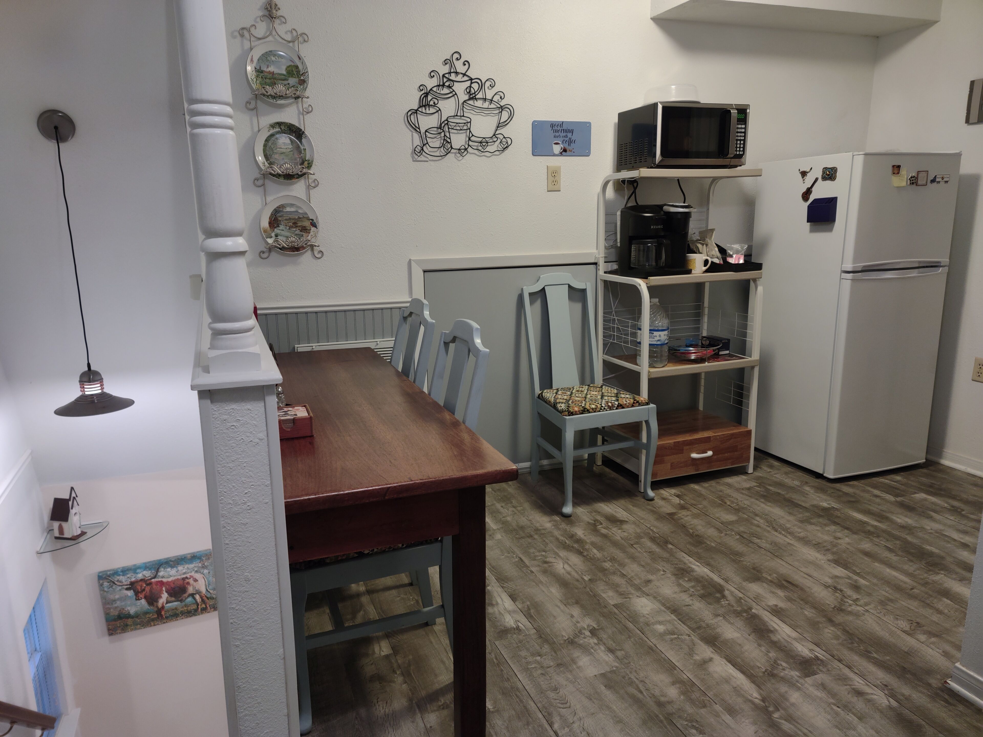 Una cocina con una mesa de madera, sillas, un microondas, un refrigerador y una estantería con utensilios de cocina.