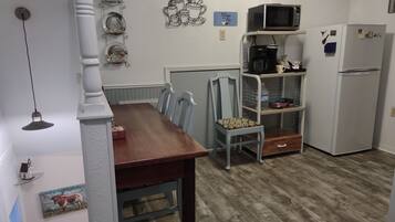 Una cocina con una mesa de madera, sillas, un microondas, un refrigerador y una estantería con utensilios de cocina.
