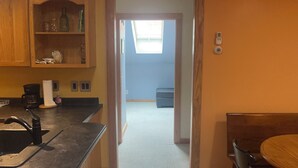 A kitchen with a sink, countertop, and wooden cabinets. There is a dining area with a table and chairs. A door leads to a hallway with a window and a blue wall. - Comfortable retreat in the woods, but close to Island attractions (Mount Desert)