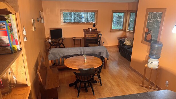 A cozy living room with a dining area, a large window, a water dispenser, and a wooden bench. - Comfortable retreat in the woods, but close to Island attractions (Mount Desert)