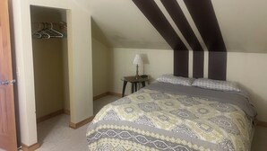 A bedroom with a sloped ceiling, a bed with patterned bedding, a bedside table with a lamp, and a coat rack. - Comfortable retreat in the woods, but close to Island attractions (Mount Desert)