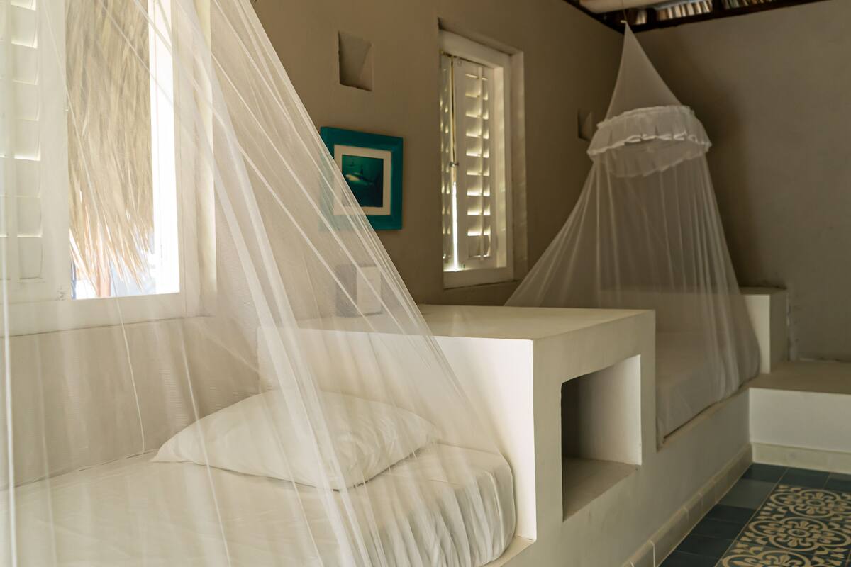 Une chambre à coucher avec un lit recouvert d’une moustiquaire, une fenêtre avec des volets et une petite table de chevet.