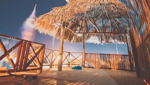 A wooden deck with a thatched roof structure, a railing, and a view of the ocean. - Kokohau Bungalows - Adults Only (Juan de Acosta)