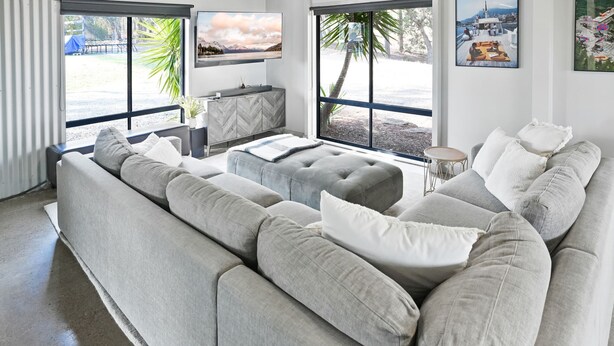 Un salon moderne avec un canapé d’angle gris, un petit pouf et un téléviseur posé sur un meuble dont le motif rappelle un chevron.