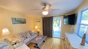 Sala de estar con sofá, sillón, mesa de centro y ventilador de techo. Hay un televisor montado en la pared y una ventana con cortinas.