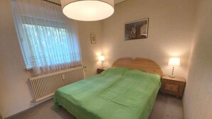 Makuuhuone, jossa on sänky, kaksi yöpöytävalaisinta, patteri, ikkuna verhoilla ja seinälle ripustettu taulu.