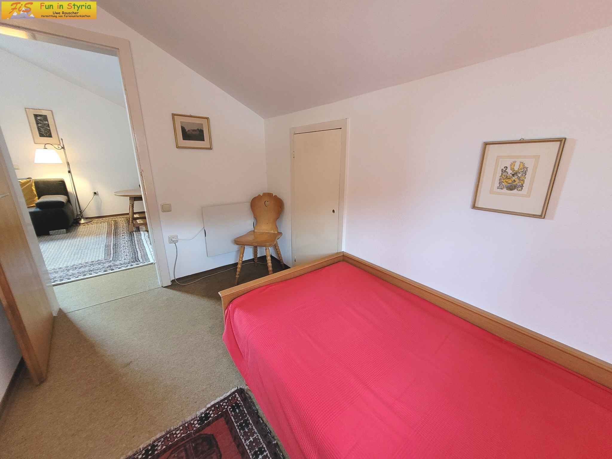 Un dormitorio con una cama roja, una silla de madera, una mesita y una puerta. Hay dos cuadros en la pared.