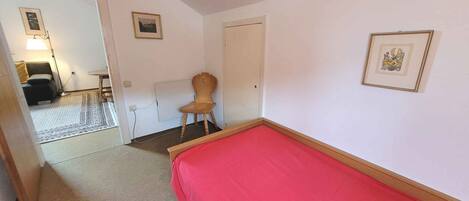 Un dormitorio con una cama roja, una silla de madera, una mesita y una puerta. Hay dos cuadros en la pared.