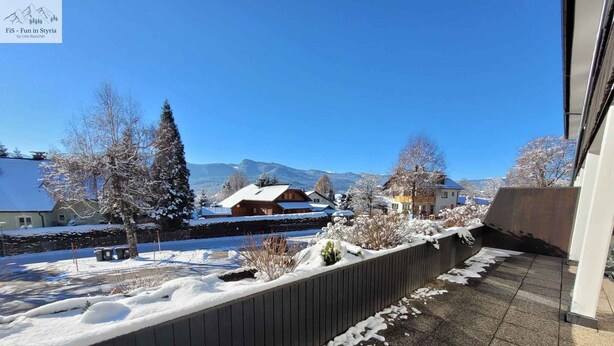 A snowy outdoor area with a wooden fence, a paved path, and houses in the background. - Holiday apartment with large balcony (Bad Mitterndorf)