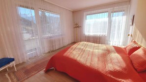 A bedroom with a large bed, two windows with sheer curtains, a floor lamp, and a blue chair. - Holiday apartment with large balcony (Bad Mitterndorf)