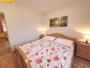 Une chambre à coucher avec un lit en bois, une literie à motifs floraux, deux lampes de chevet, un tableau représentant une forêt et un plancher en bois.