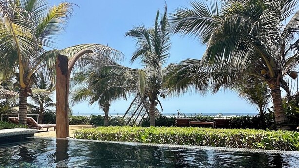 Una piscina cristalina con fondo de baldosas, rodeada de palmeras y un cielo despejado.