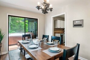 Uma sala de jantar com uma mesa de madeira posta para quatro pessoas, um candelabro e vista para uma floresta através de uma porta de vidro.