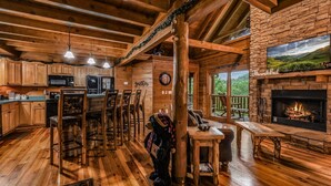A rustic cabin interior with wooden floors, a stone fireplace, a dining area, and a kitchen with wooden cabinets and bar stools.