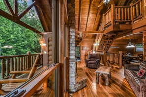 A wooden cabin interior with a stone fireplace, wooden furniture, and a balcony overlooking greenery.