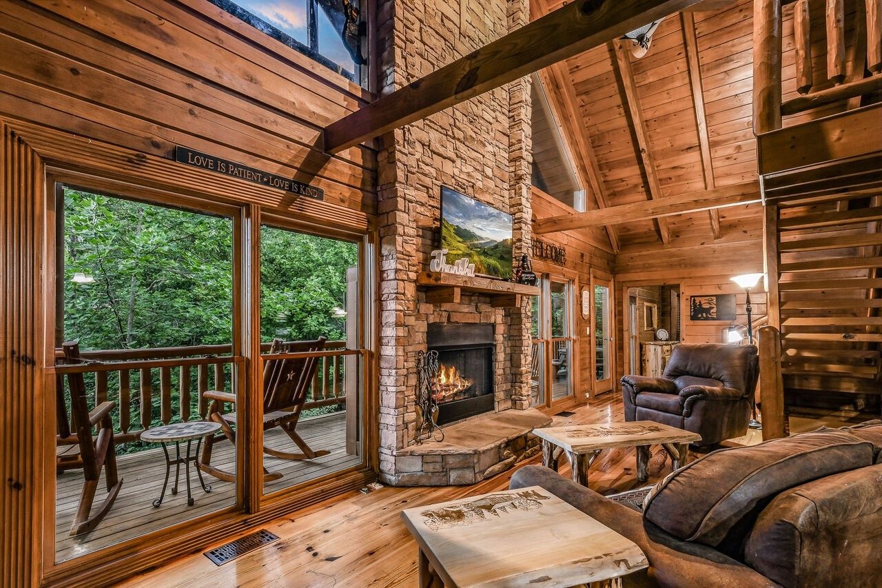 A wooden cabin interior with a stone fireplace, wooden deck, and a large window overlooking greenery.