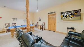 Une pièce avec une table de billard, des fauteuils en cuir, une porte en bois et un téléviseur fixé au mur qui diffuse un événement sportif.