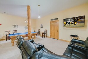 Een woonkamer met een pooltafel, leren fauteuils, een houten deur en een televisie waarop een voetbalwedstrijd wordt vertoond.