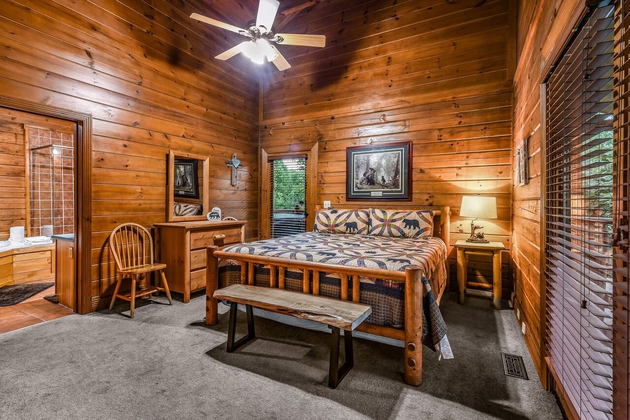 A wooden cabin bedroom with a bed, a desk, a chair, a bench, and a ceiling fan.