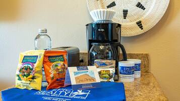 Una encimera de cocina con una cafetera, una botella, bolsas de aperitivos hawaianos, una toalla azul y un reloj de pared.