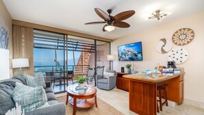 A modern living room with a large sofa, a coffee table, and a kitchen area with a bar. There is a ceiling fan, a TV, and a view of the ocean through large windows. - Kuleana 613 1 Bdrm 1 Bedroom Condo by RedAwning (Lahaina)