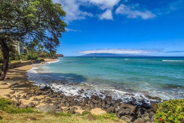 A beach with clear blue water, a rocky shoreline, and a tree-lined path. - Kuleana 613 1 Bdrm 1 Bedroom Condo by RedAwning (Lahaina)