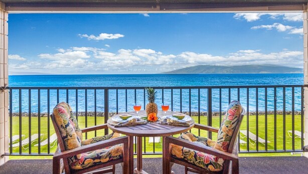 A balcony with a table and chairs, overlooking a clear blue sea and a distant mountain. - Kaleialoha 209 1 Bedroom Condo by RedAwning (Lahaina)