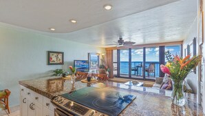 A kitchen with a large island, a dining area with chairs, and a view of the ocean through large windows. - Kaleialoha 209 1 Bedroom Condo by RedAwning (Lahaina)