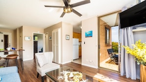 A modern living room with a glass coffee table, a ceiling fan, and a view of the kitchen and outdoor patio. - Kahana Manor 418 1 Bedroom Condo by RedAwning (Lahaina)